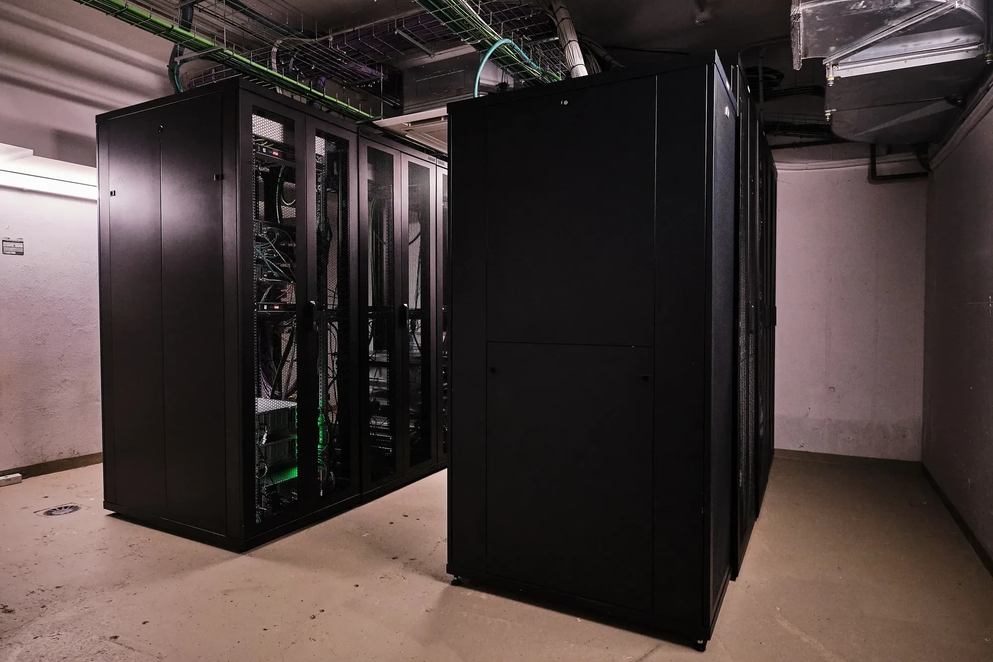Sala de servidores no soto: armarios rack negros, un deles con portas de malla que deixan ver equipos iluminados en verde, baixo bandexas de cableado e condutos de climatización.