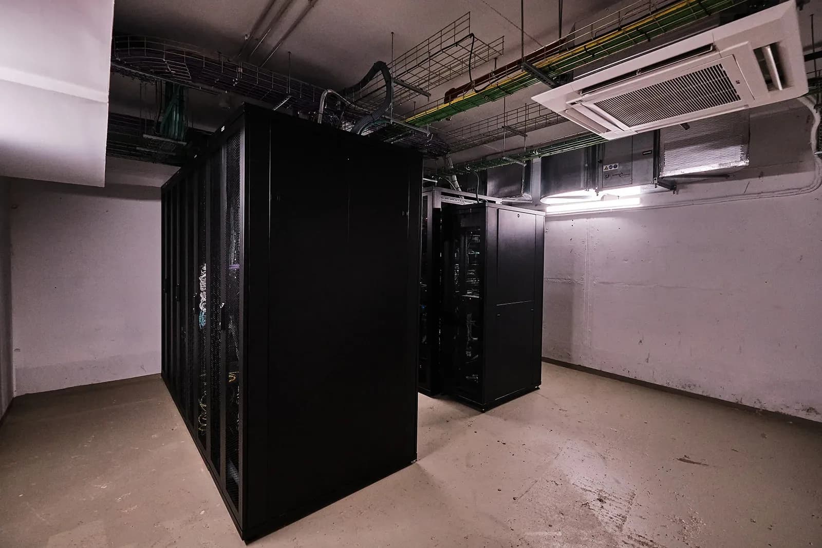 Sala de servidores no soto: dous bloques de armarios rack negros baixo bandexas de cableado e unha unidade de climatización de teito.