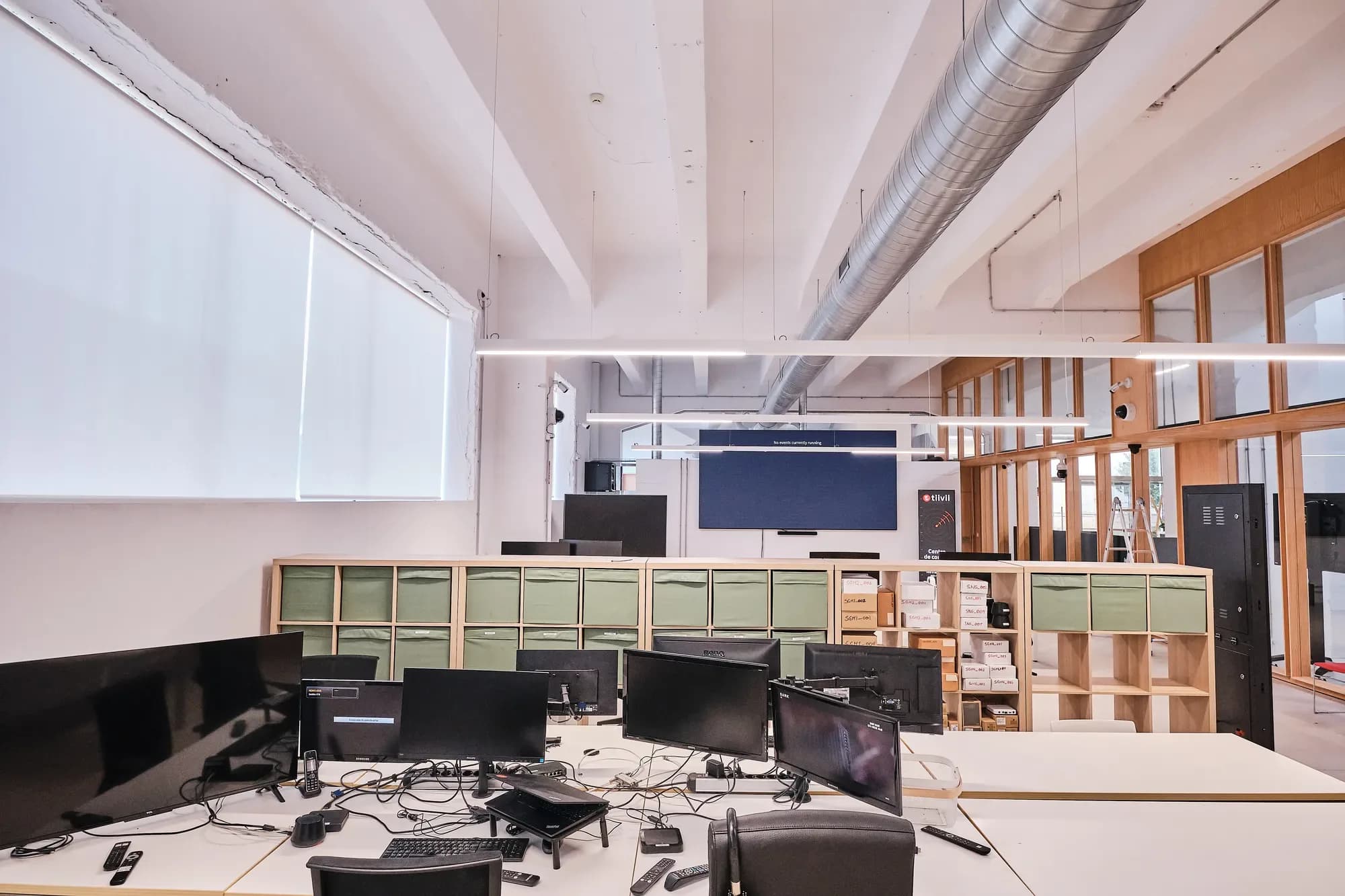 Laboratorio de medios: mesas con varios monitores, descodificadores, mandos a distancia e cableado; detrás, estanterías con caixas e unha gran pantalla LED na parede do fondo.