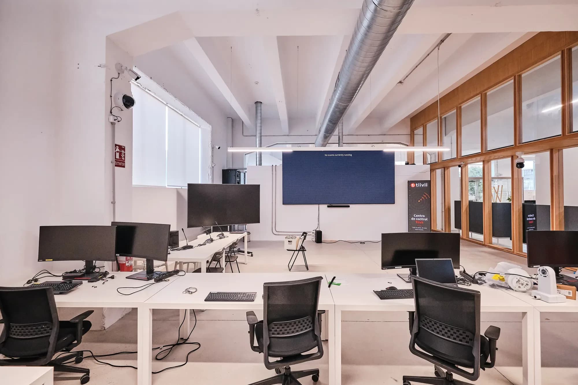 Laboratorio de medios: fila de postos con monitores e cadeiras de oficina; ao fondo, unha gran pantalla LED e un roll-up de Tiivii xunto á cristaleira de madeira.