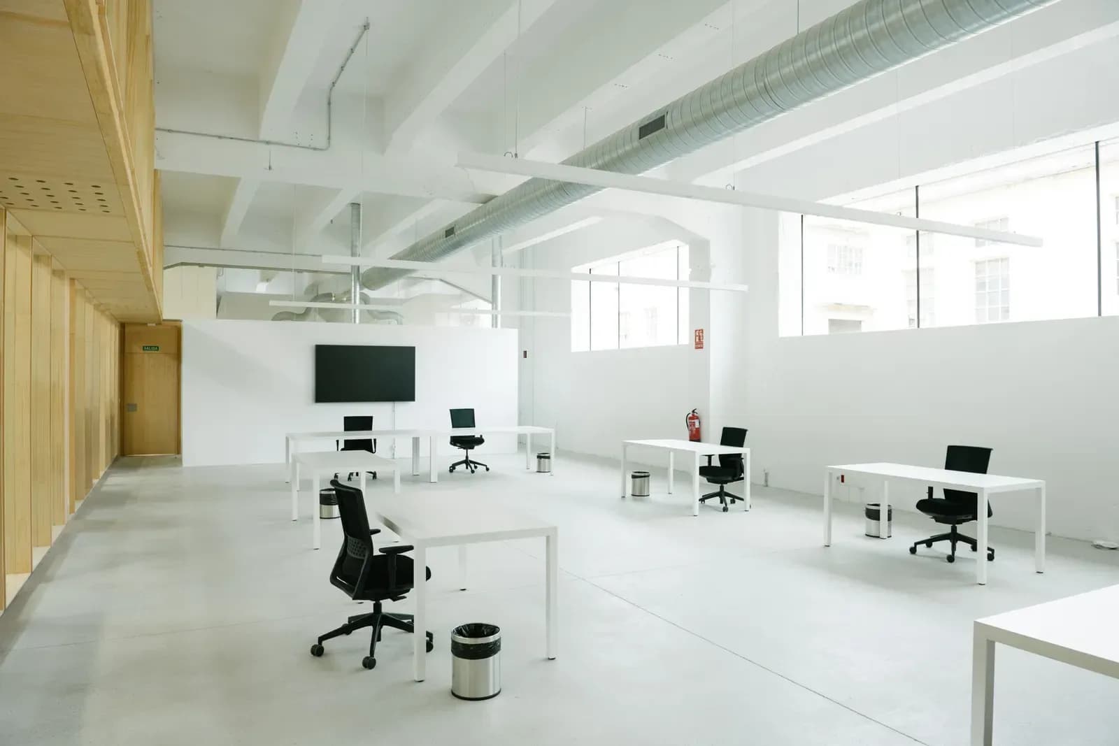 Oficina diáfana rehabilitada, con mesas de traballo e monitor sobre chan continuo claro.