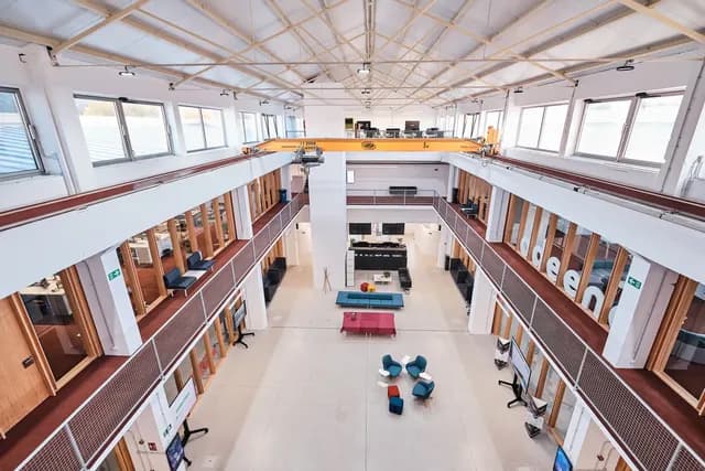 Vista xeral do atrio desde o alto: pasarelas laterais con oficinas acristaladas, grupos de butacas e sofás sobre o chan de formigón e a grúa amarela ao fondo.